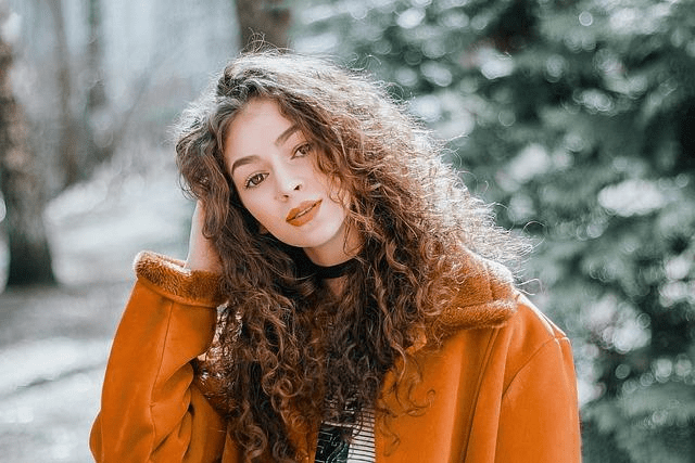 A young woman with curly hair wears an orange jacket to pose in front of the camera.